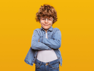 Smart little ginger nerd boy with curly hair wearing denim outfit and round glasses standing with arms crossed against yellow background and looking at camera