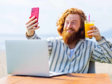 young redhaired ginger bearded man working outdoors in sea cafe with laptop an summer day.