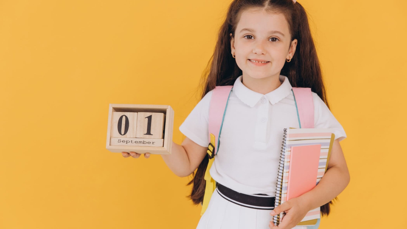 Portrait of first grader little girl looking at camera and hold wooden calendar set on 1 september isolated yellow background. Back to school