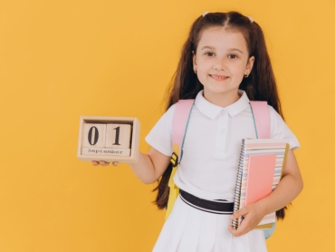 Portrait of first grader little girl looking at camera and hold wooden calendar set on 1 september isolated yellow background. Back to school