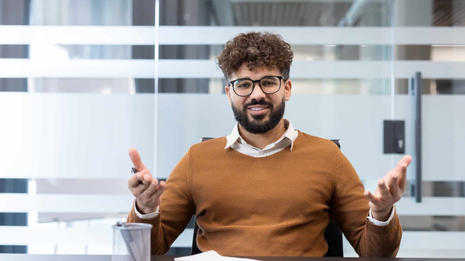 Young smiling businessman in glasses and beard sitting at a conference table, actively communicating and sharing ideas during a corporate discussion or consultation in a contemporary office setting