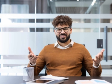 Young smiling businessman in glasses and beard sitting at a conference table, actively communicating and sharing ideas during a corporate discussion or consultation in a contemporary office setting