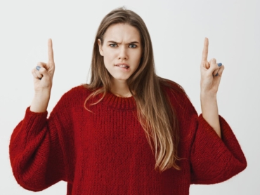 Hesitating attractive caucasian female model in trendy red loose sweater, raising index fingers and pointing up, biting lip, frowning, being intense and worried, being doubtful and making decision. Copy space